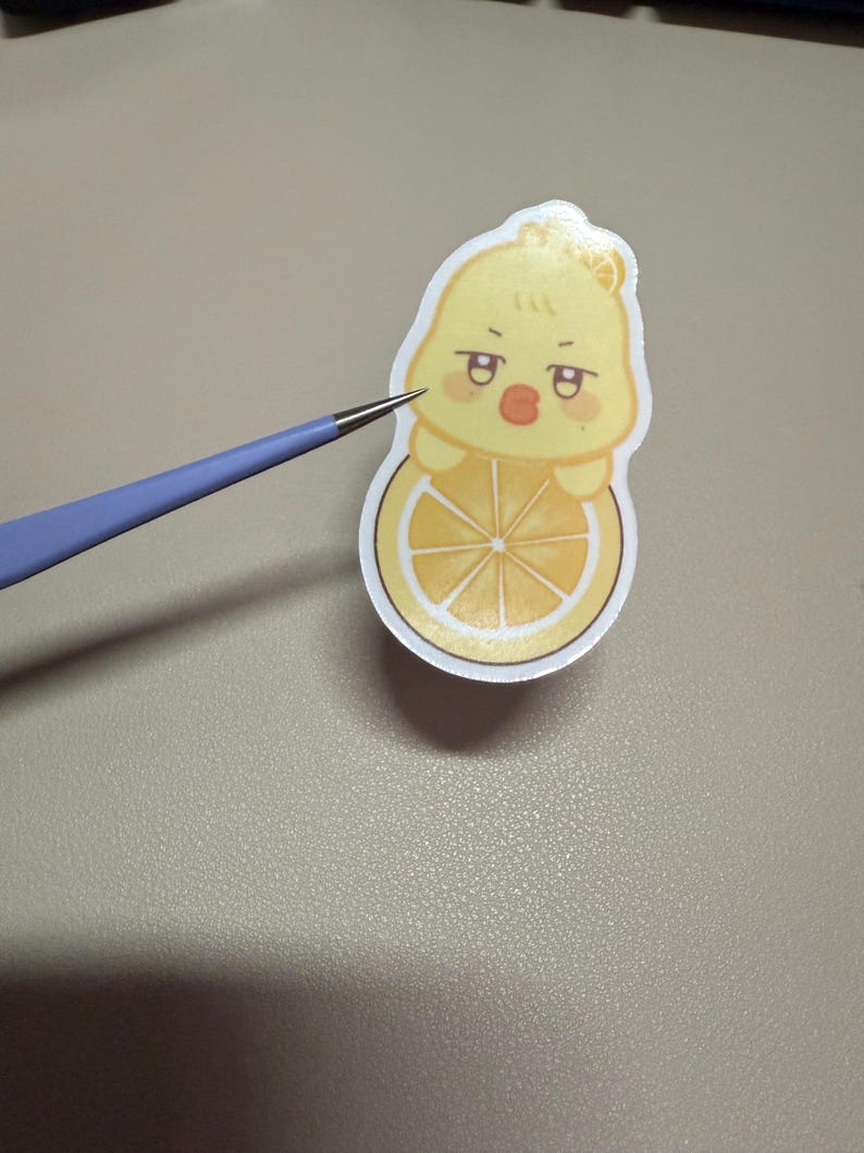 Op de afbeelding: Een gele sticker met een cartoon eend karakter met een sinaasappelschijf. De eend heeft een chagrijnige uitdrukking en houdt de sinaasappelschijf vast. De sticker wordt vastgehouden door een blauw en zilverkleurig gereedschap.