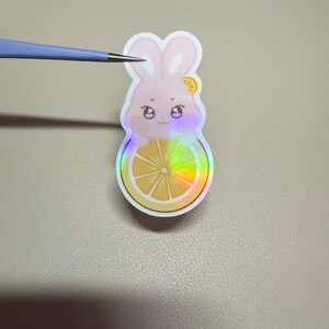 Op de afbeelding: Een holografische sticker met een roze konijn met een citroen schijfje op zijn hoofd, zittend op een citroen schijfje. De sticker heeft een regenboog iriserend effect. Het konijn heeft een schattige uitdrukking.