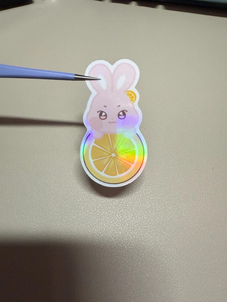 Op de afbeelding: Een holografische sticker met een roze konijn met een citroen schijfje op zijn hoofd, zittend op een citroen schijfje. De sticker heeft een regenboog iriserend effect. Het konijn heeft een schattige uitdrukking.