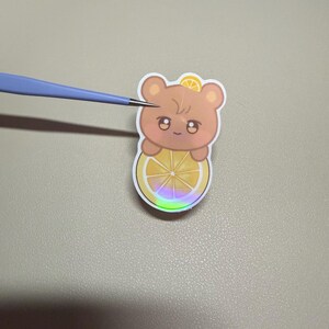 Op de afbeelding: Een sticker met een cartoonbeer met een citroenschijfje. De beer is lichtbruin met een klein citroenschijfje op zijn hoofd. De beer houdt een grotere citroenschijf vast met een regenboog holografisch effect.