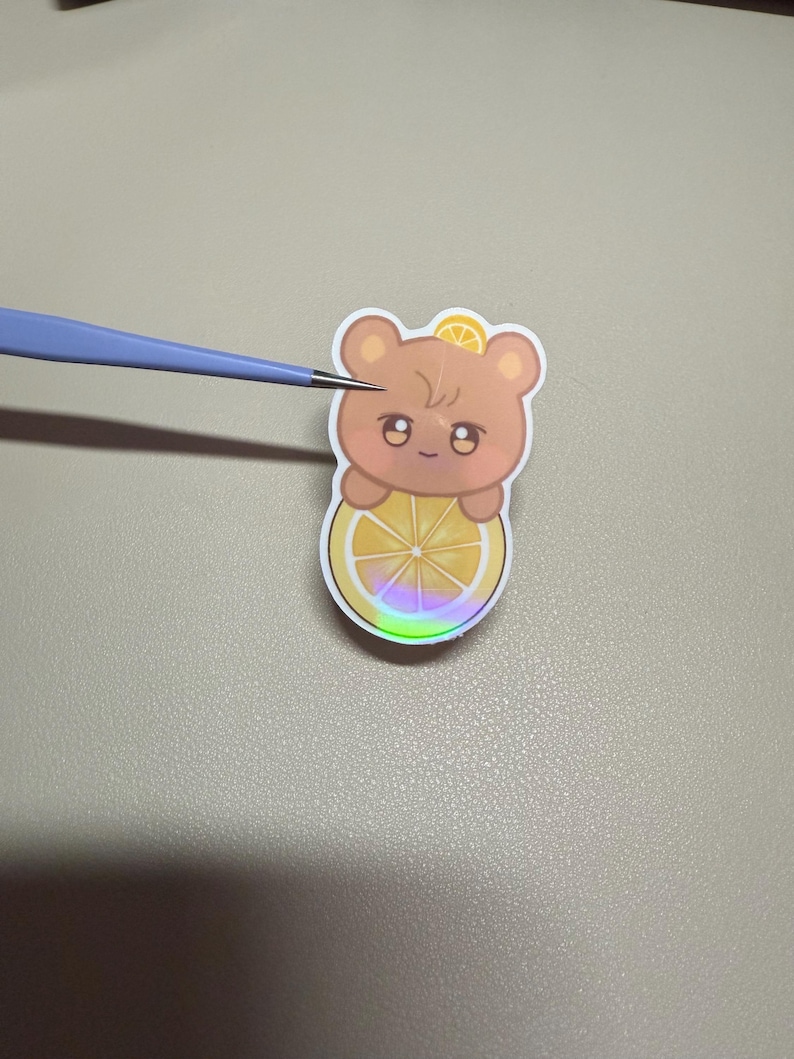 Op de afbeelding: Een sticker met een cartoonbeer met een citroenschijfje. De beer is lichtbruin met een klein citroenschijfje op zijn hoofd. De beer houdt een grotere citroenschijf vast met een regenboog holografisch effect.