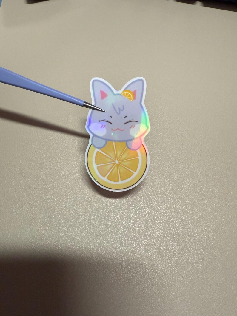 Op de afbeelding: Een holografische sticker met een lachend kattenpersonage op een citroen. De kat is lichtgrijs met een klein sinaasappelschijfje op zijn hoofd. De sticker heeft een regenboogglans.