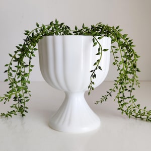 Op de afbeelding: Een witte, gegroefde voetplanter met een brede kom en een afgeronde basis. Hangende groene plantenstengels vallen over de zijkanten. De planter is gemaakt van een glanzend, ondoorzichtig materiaal en de plant voegt een natuurlijk tintje toe.