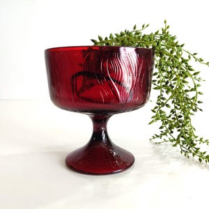 May include: A ruby red glass pedestal bowl with a textured, bark-like pattern. The bowl has a wide, round top and a short, flared stem. A sprig of green foliage is placed behind the bowl.
