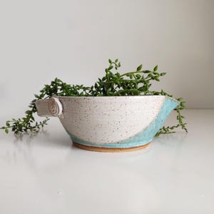 May include: A handmade ceramic bowl with a white speckled finish and a turquoise accent. The bowl features a unique handle and is filled with green trailing plants. The bowl is a decorative piece for the home.