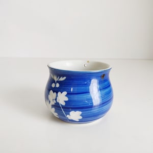 Op de afbeelding: Een kleine, ronde blauwe keramische kom met witte bloemmotieven. De kom heeft een wit interieur en een gestreepte blauwe buitenkant. Twee kleine gaatjes zijn zichtbaar bij de rand.