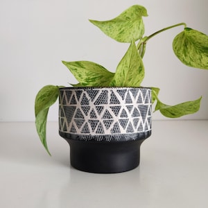 May include: A black and white geometric patterned ceramic planter with a green plant inside.