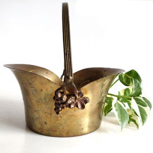 May include: Vintage brass basket with a handle and decorative grape cluster detail. The basket has a flared rim and a sturdy handle. The brass has a warm, aged patina. A decorative piece for storage or display.