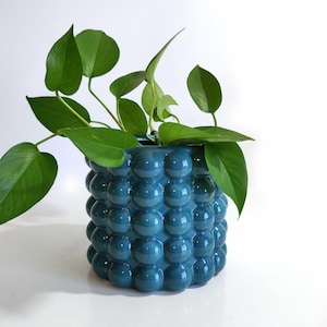 May include: A cylindrical teal ceramic planter featuring a raised bubble texture across its exterior surface. A vibrant green pothos plant with heart-shaped leaves grows out of the top of the pot, with several stems cascading over the sides.