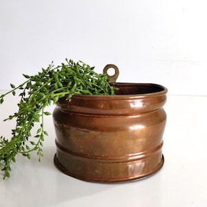 Peut inclure: Une jardinière murale ronde en cuivre avec une finition métallique polie et une patte de fixation circulaire unique sur le dessus. Le récipient présente un design nervuré horizontal sur le corps et contient une plante verte retombante.