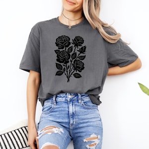 May include: A charcoal gray t-shirt featuring a black floral design of roses and leaves. The graphic is centered on the front of the shirt. The model is wearing blue jeans.