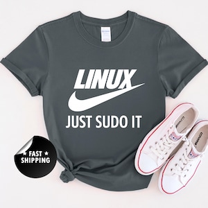 May include: Dark gray t-shirt with the text "LINUX" above a white swoosh and the words "JUST SUDO IT" in white. A pair of white sneakers with red accents are next to the shirt.
