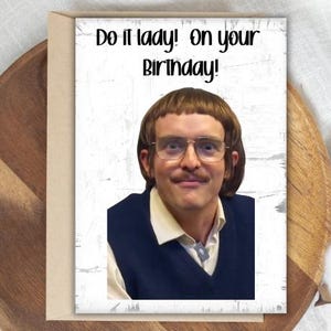 May include: A birthday card with a photo of a man wearing glasses and a mustache. The card has the text "Do it lady! On your Birthday!" in black font. The card is on a wooden tray.