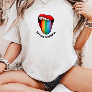 Könnte beinhalten: Weißes Kurzarm-T-Shirt mit einer Grafik von roten Lippen und einer Regenbogenzunge. Der Text "THE FUTUR IS INCLUSIVE" ist darunter in Schwarz gedruckt. Das T-Shirt hat einen lässigen Stil.