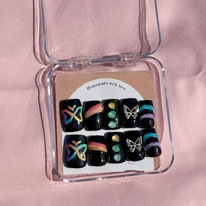 May include: A set of black press-on nails with various designs, including butterflies, rainbow accents, and gemstones, displayed in a clear plastic case. The text "Homemade with love" is visible on the packaging.