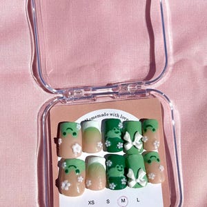May include: A set of press-on nails in a clear plastic case. The nails feature a variety of designs, including green and beige with white flower accents and bows. The text "Homemade with love" is visible on the label, along with sizes XS, S, M, and L.