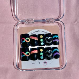 Könnte beinhalten: Ein Set schwarzer Kunstnägel mit verschiedenen Designs, darunter Regenbogen-Unendlichkeitszeichen, farbige Streifen und Edelsteinakzente. Die Nägel werden in einer transparenten Kunststoffbox mit den Größen XS, S, M und L präsentiert.