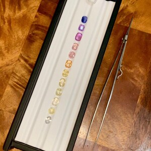 May include: A tray with a black frame holds a row of colorful gemstones, ranging from blue to yellow. A pair of silver tweezers rests beside the tray. The gemstones are oval-shaped and set against a white background.
