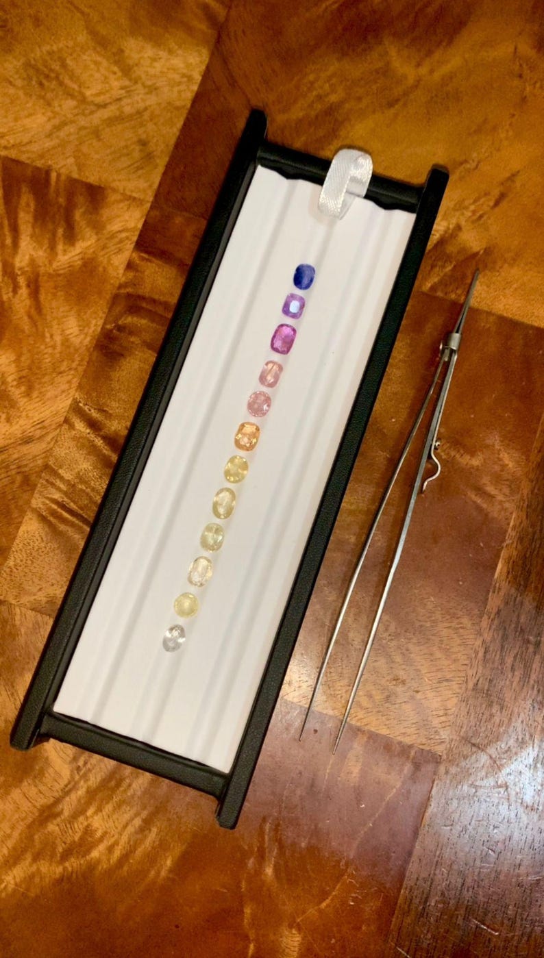 May include: A tray with a black frame holds a row of colorful gemstones, ranging from blue to yellow. A pair of silver tweezers rests beside the tray. The gemstones are oval-shaped and set against a white background.