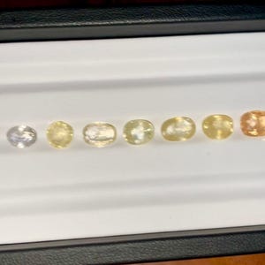 May include: A collection of oval-cut gemstones in varying shades of yellow and orange, displayed in a white tray. The gemstones range from pale yellow to a deep orange hue, showcasing a gradient of colors. The tray is set against a dark background.