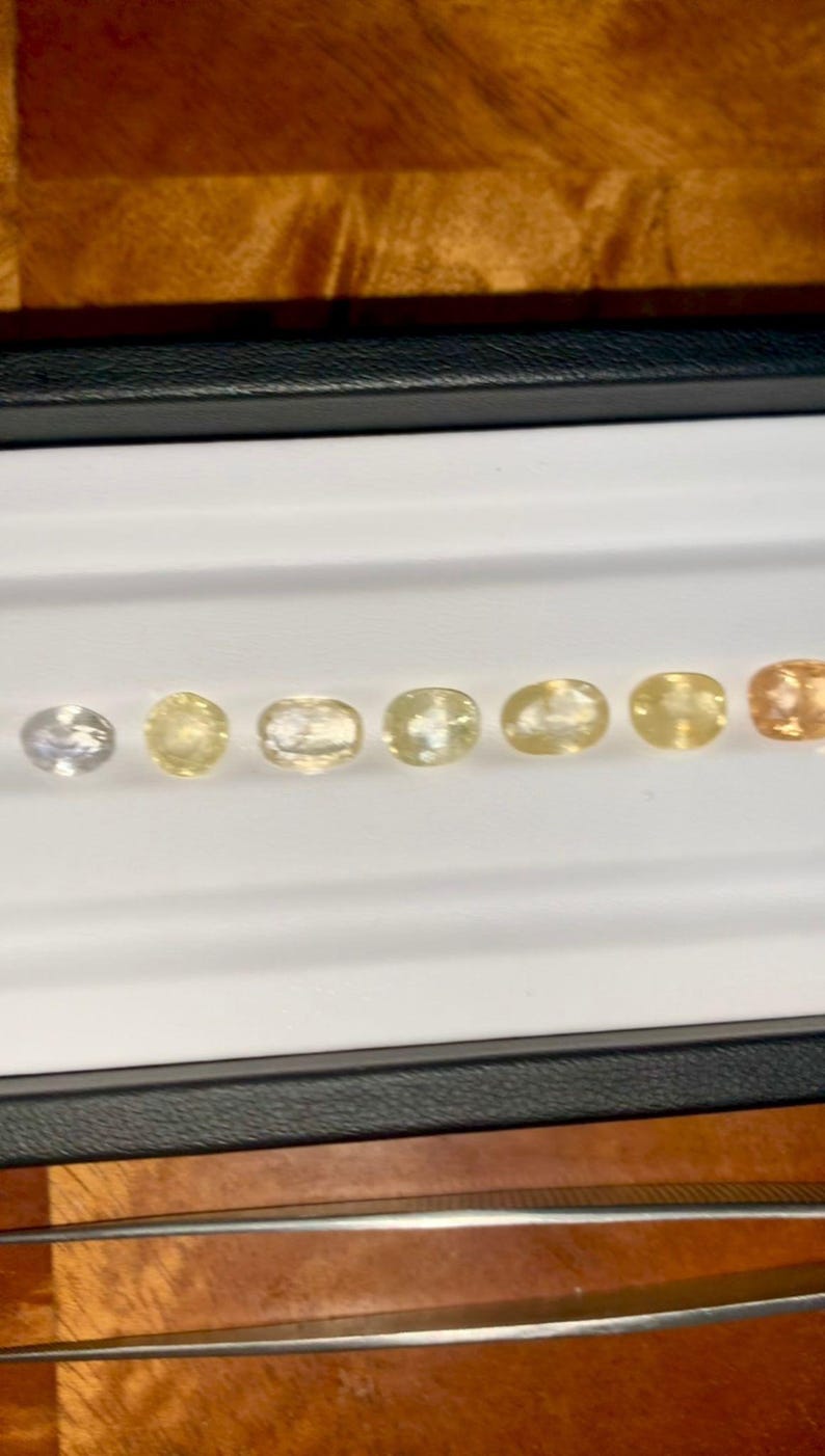 May include: A collection of oval-cut gemstones in varying shades of yellow and orange, displayed in a white tray. The gemstones range from pale yellow to a deep orange hue, showcasing a gradient of colors. The tray is set against a dark background.