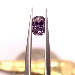 May include: A radiant-cut, purplish-pink gemstone held by silver tweezers. The gem has a rectangular shape with cut corners and facets that reflect light. The background is white, with a hint of gold at the bottom.