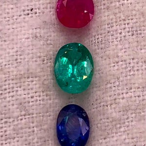 May include: Three oval-cut gemstones arranged vertically. The top stone is a vibrant, deep pinkish-red. The middle stone is a bright, translucent green with visible internal inclusions. The bottom stone is a dark, saturated royal blue.