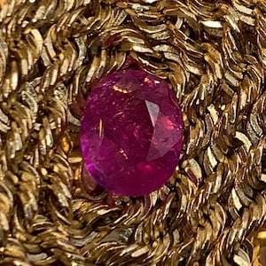 May include: A deep pink, oval-cut gemstone sits on a gold chain with a woven pattern. The gemstone's facets catch the light, creating a sparkling effect. The image evokes a sense of luxury and is likely a piece of jewellery.