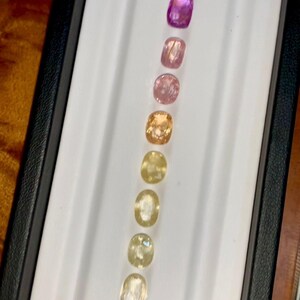 May include: A collection of cushion-cut gemstones in a range of colors, from deep blue to pale yellow. The gems are arranged in a row inside a white tray with a black border, showcasing their varying hues and clarity.
