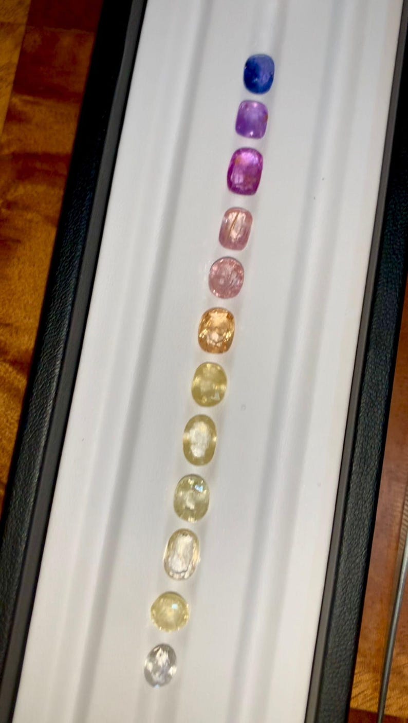 May include: A collection of cushion-cut gemstones in a range of colors, from deep blue to pale yellow. The gems are arranged in a row inside a white tray with a black border, showcasing their varying hues and clarity.