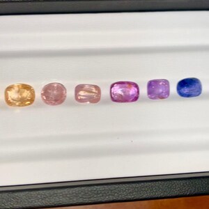 May include: A collection of six gemstones in a variety of colors, including peach, pink, and purple, arranged in a row. The gemstones are cushion-cut and displayed in a white tray with a black border. A pair of tweezers is visible.