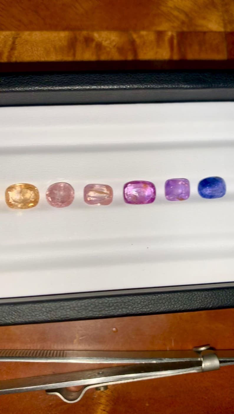 May include: A collection of six gemstones in a variety of colors, including peach, pink, and purple, arranged in a row. The gemstones are cushion-cut and displayed in a white tray with a black border. A pair of tweezers is visible.