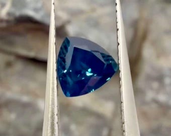 May include: A deep blue, trillion-cut gemstone held by a pair of silver-toned metal tweezers. The stone features a faceted surface that reflects light, displaying various shades of blue throughout its triangular shape and polished edges.
