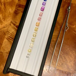 May include: A collection of gemstones in a black display case with a white interior. The gemstones are arranged in a row, showcasing a gradient of colors from blue to yellow. A pair of tweezers rests beside the case.