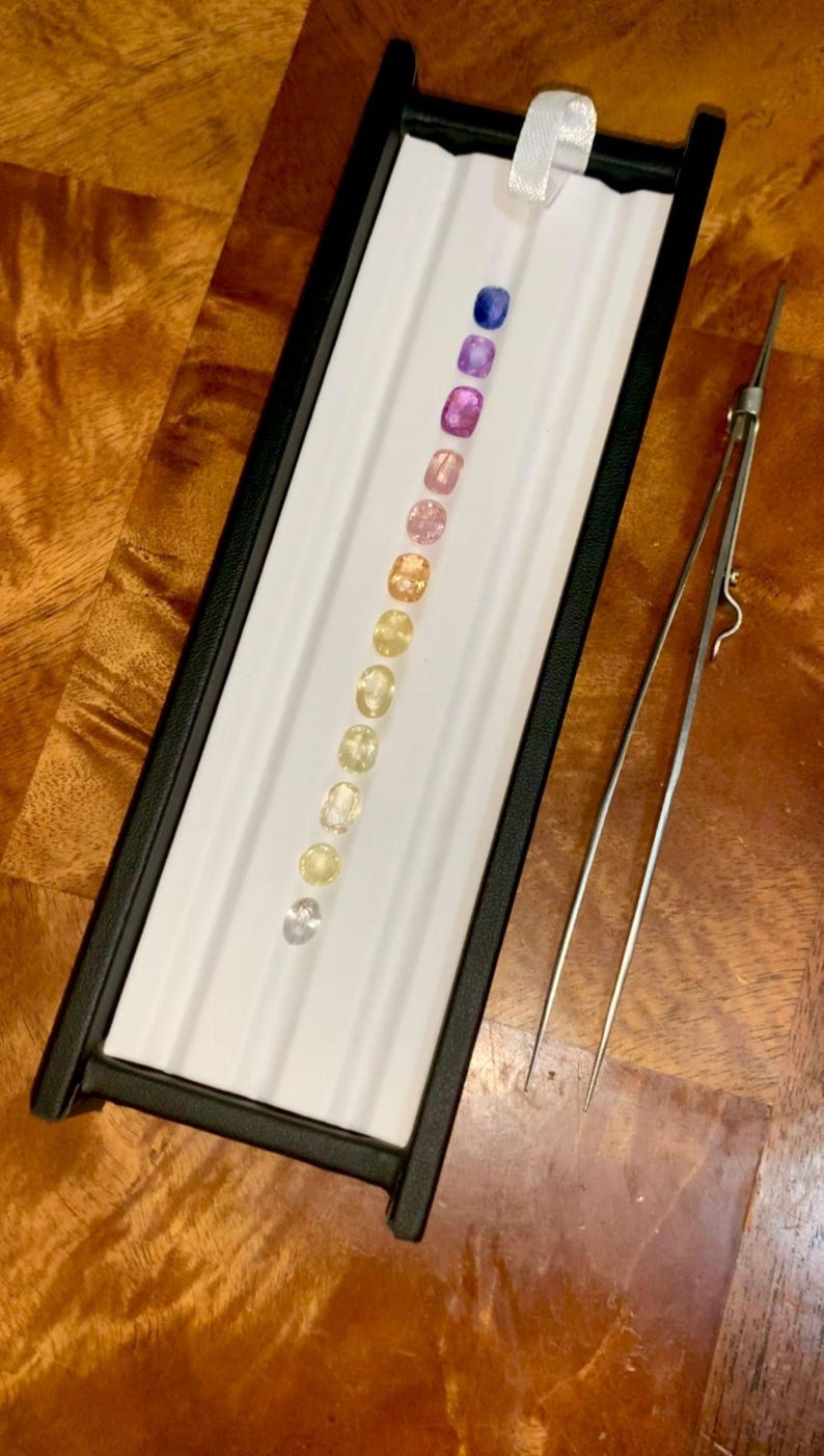 May include: A collection of gemstones in a black display case with a white interior. The gemstones are arranged in a row, showcasing a gradient of colors from blue to yellow. A pair of tweezers rests beside the case.