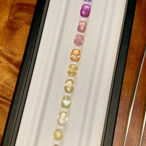 May include: A collection of gemstones in a variety of colors, including blue, purple, pink, and yellow, are displayed in a black box with a white interior. The gemstones are oval-shaped and arranged in a vertical line. Tweezers are visible on the right.