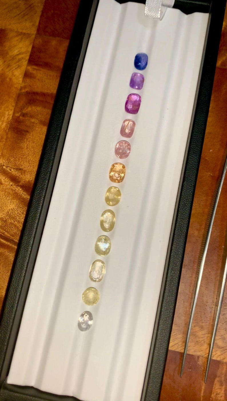 May include: A collection of gemstones in a variety of colors, including blue, purple, pink, and yellow, are displayed in a black box with a white interior. The gemstones are oval-shaped and arranged in a vertical line. Tweezers are visible on the right.