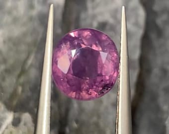 May include: A faceted, oval-shaped gemstone in shades of pink and purple is held by silver tweezers. The gem has a brilliant cut, reflecting light. The gemstone is a vibrant, eye-catching piece, perfect for jewelry making.