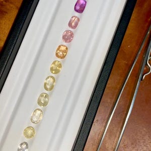 May include: A collection of oval-cut gemstones in a range of colors, from clear to deep blue, arranged in a white tray. A pair of tweezers is positioned to the right. The gemstones are likely sapphires or similar precious stones.