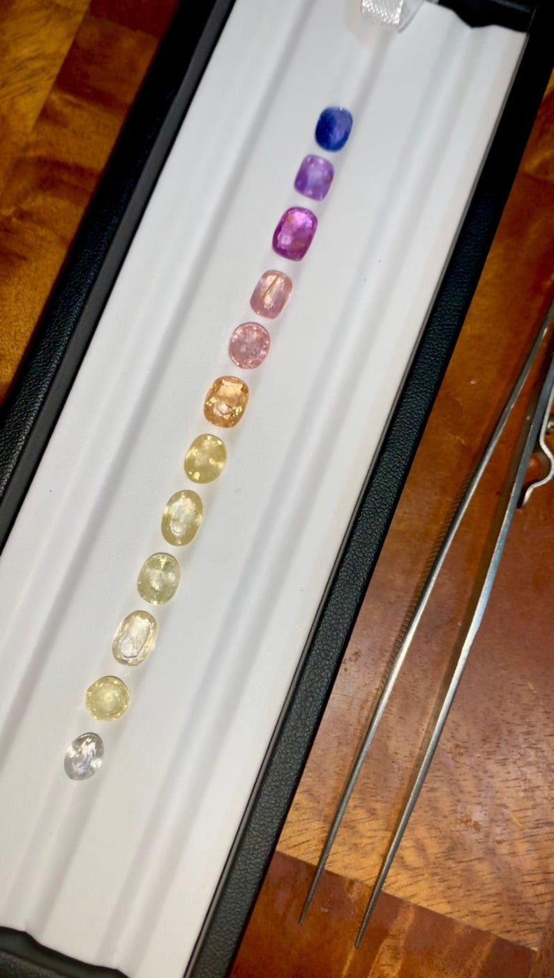 May include: A collection of oval-cut gemstones in a range of colors, from clear to deep blue, arranged in a white tray. A pair of tweezers is positioned to the right. The gemstones are likely sapphires or similar precious stones.