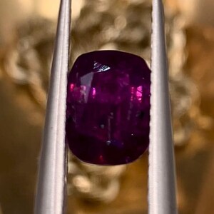 以下が含まれることがあります： 銀色のピンセットで保持された、ファセット加工が施された濃いマゼンタ色の宝石。宝石は丸みを帯びた長方形の形状をしており、光を反射し、内部構造を示しています。背景はぼやけた金色です。