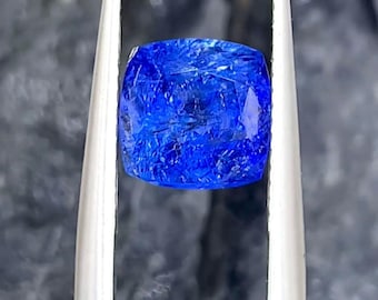 May include: A vibrant, cushion-cut blue gemstone held by silver-toned metal tweezers. The stone features a faceted surface with visible internal inclusions and a deep, saturated cobalt blue color that reflects light across its polished top surface.