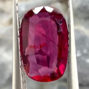 May include: A faceted, oval-shaped ruby gemstone held by silver tweezers. The ruby is a deep, saturated red color with a high degree of clarity. The gemstone is cut to maximize light reflection and brilliance, showcasing its natural beauty.
