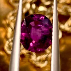 以下が含まれることがあります： 鮮やかなオーバルカットの濃いマゼンタ色の宝石が、ぼやけた金色の背景に対して銀色のピンセットで保持されています。宝石のファセットは光を反射し、その豊かな色と透明度を示しています。この画像は宝石の美しさを際立たせています。