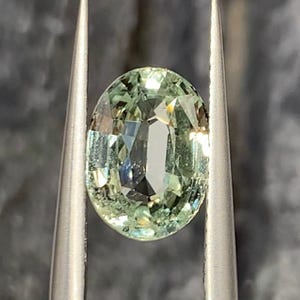 May include: An oval-cut, pale green gemstone held by silver tweezers. The gem has a faceted surface that reflects light, creating a sparkling effect. The stone's color is a soft, muted green.