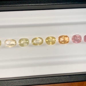 May include: A collection of oval-cut gemstones in a range of colors, from pale yellow to pink, displayed in a black tray with a white interior. The gemstones are arranged in a row, showcasing their varying hues and clarity. A pair of tweezers is visible in the foreground.