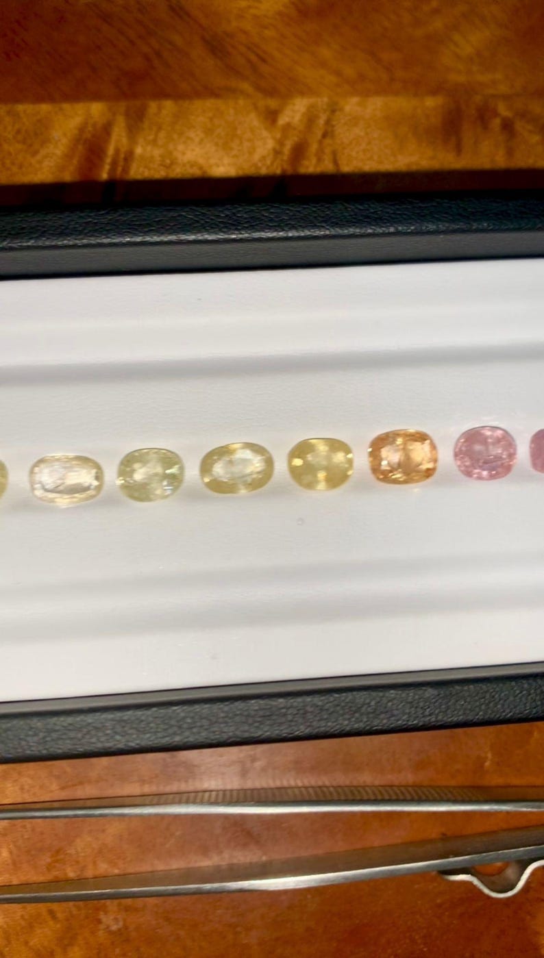 May include: A collection of oval-cut gemstones in a range of colors, from pale yellow to pink, displayed in a black tray with a white interior. The gemstones are arranged in a row, showcasing their varying hues and clarity. A pair of tweezers is visible in the foreground.