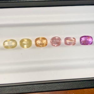 May include: A selection of six gemstones in a range of colors, from pale yellow to deep purple, displayed in a white tray. The gemstones are oval-shaped and appear to be polished. The tray is set against a dark background.