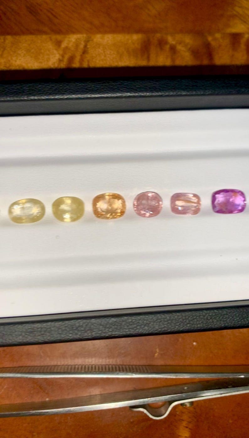 May include: A selection of six gemstones in a range of colors, from pale yellow to deep purple, displayed in a white tray. The gemstones are oval-shaped and appear to be polished. The tray is set against a dark background.