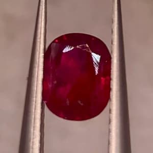 May include: A vibrant, oval-cut ruby gemstone held by silver tweezers. The ruby displays a deep red hue with internal reflections, showcasing its clarity and brilliance. The tweezers provide a contrasting metallic backdrop, highlighting the gem's colour and shape.